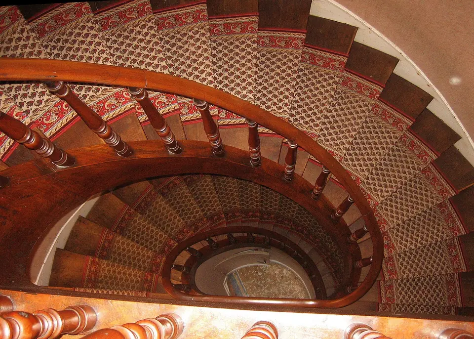 Escalier hélicoïdal en bois verni vu d'en haut, avec rampe moulurée, balustres tournés et marches couvertes d'un tapis à motifs rouges.