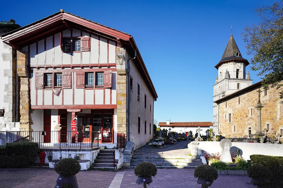 Maison labourdine à colombages et volets rouges à Ainhoa, à côté du clocher de l'église, sous un ciel bleu.